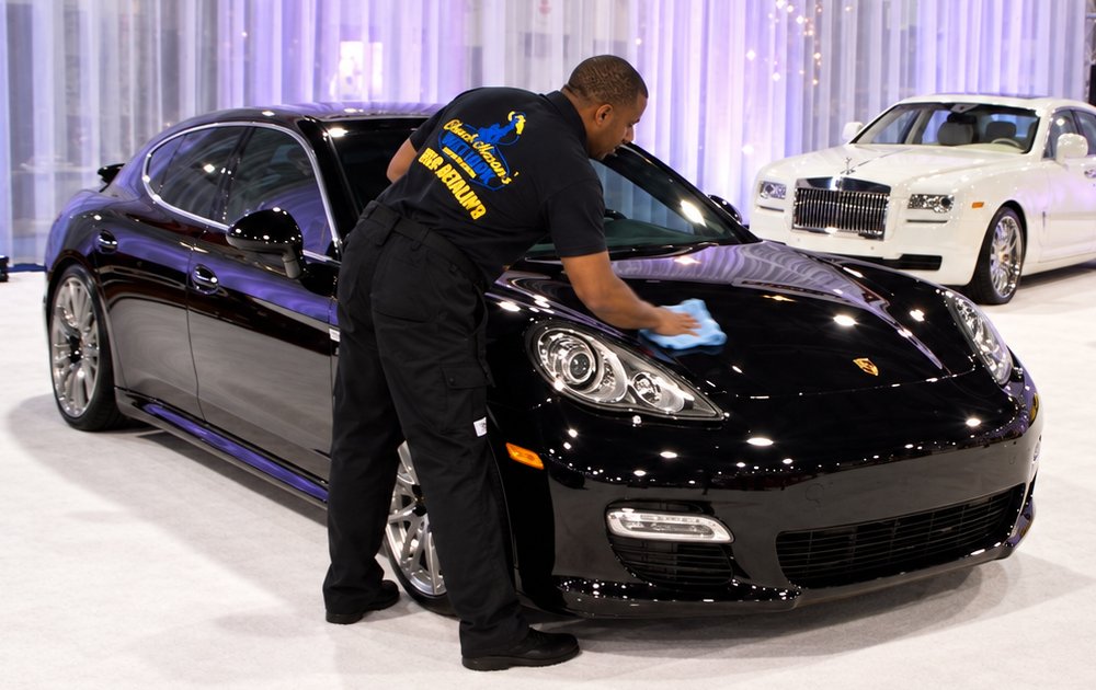 Drying the hood of a black sedan by hand on a show floor display, wearing a Chuck Mason's Wet Look shirt